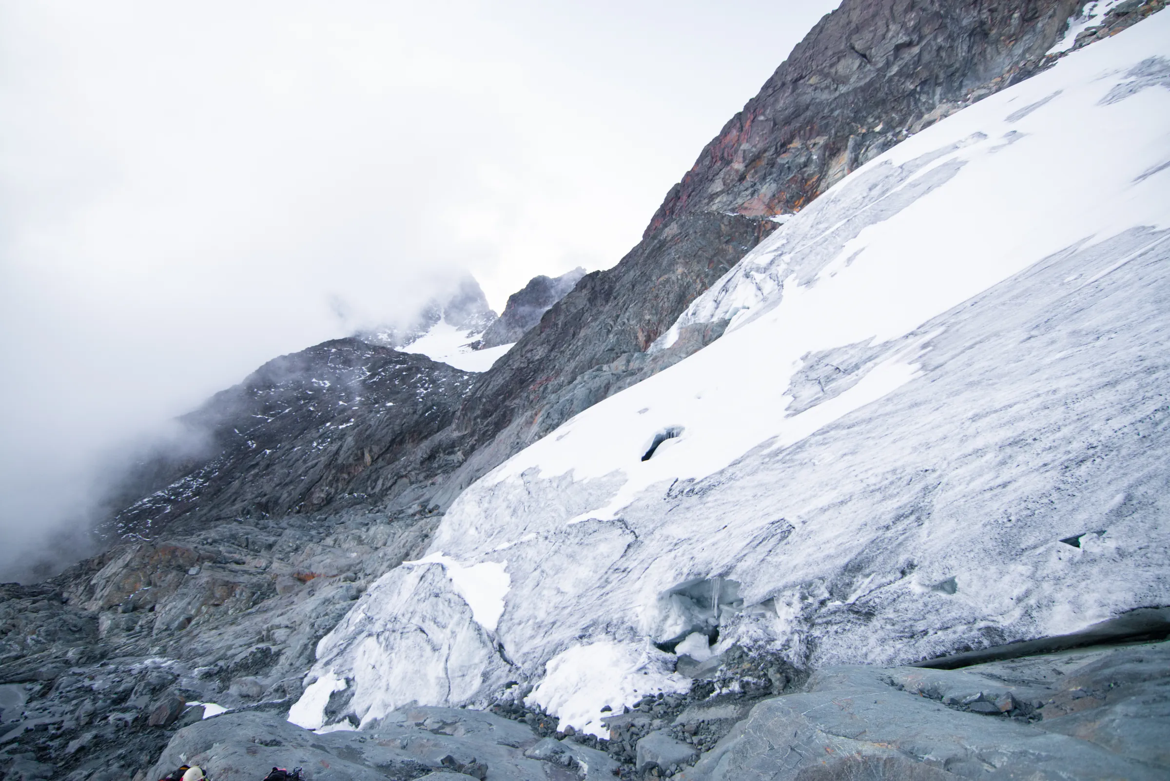 The snout of a Rwenzori glacier emerging from mist below a dark rock band, with climbers small at the lower edge
