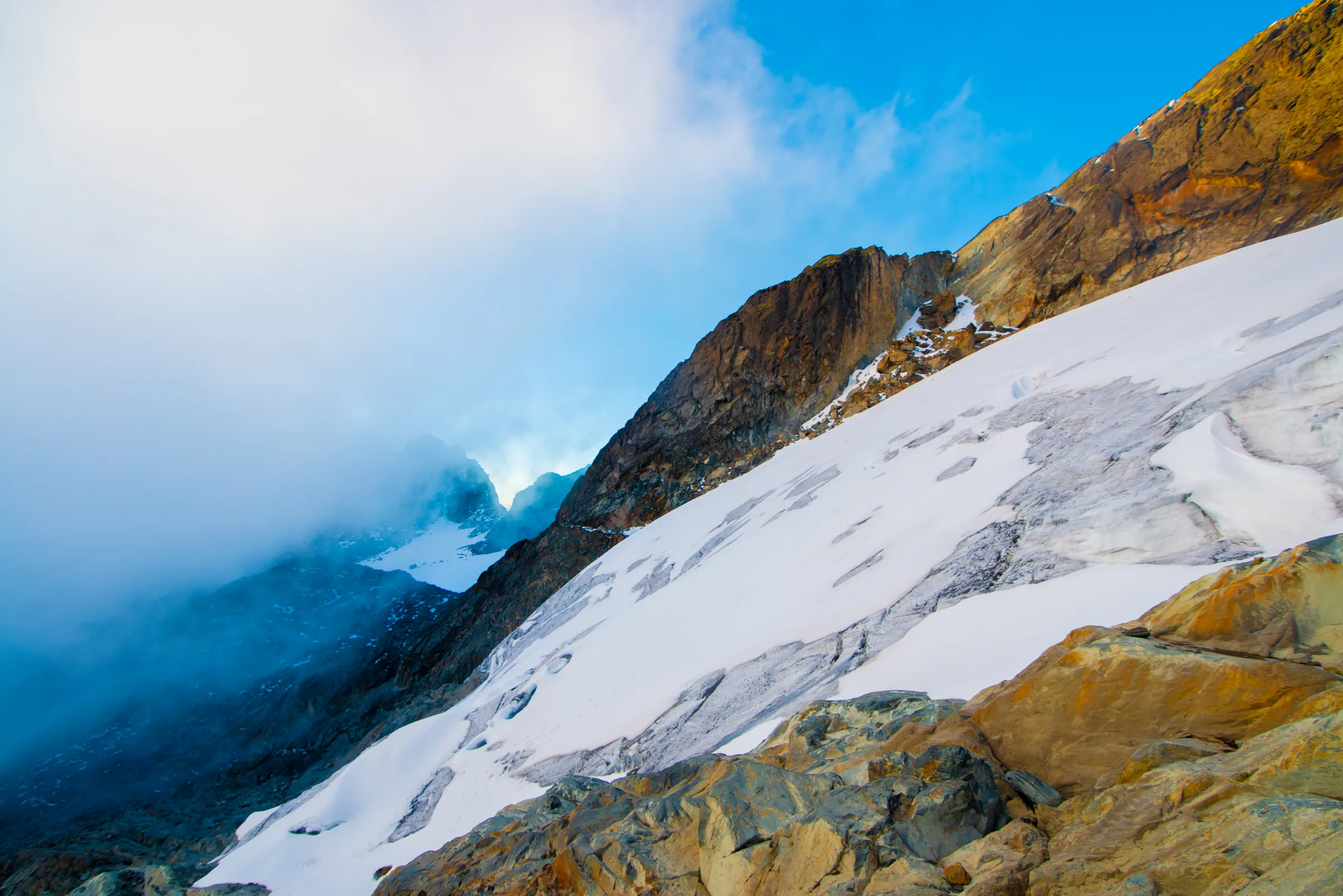 A broad snowfield sweeping below a sunlit rock ridge on the Rwenzori, cloud pressing in from the left