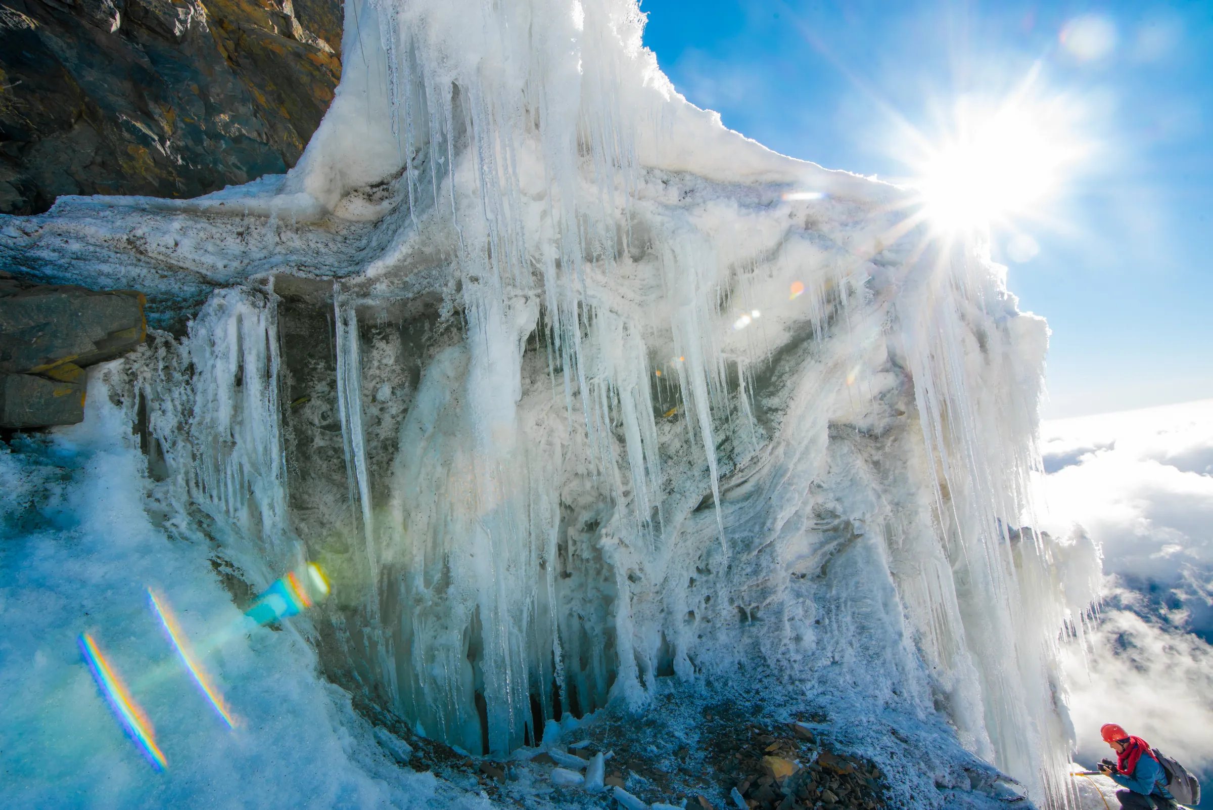 Sun bursting over a Rwenzori glacier wall hung with long icicles, a climber small at the lower right for scale